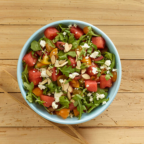 Watermelon, arugula, and tomatoes, garnished with almonds and feta, served in a blue bowl atop a wooden table.