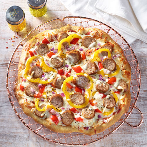 A pizza made from golden brown crust, melted cheese, sweet turkey sausage, bell peppers, and red onion on a copper rack, served with a side of olive oil.