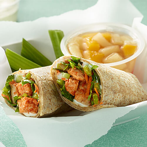 Buffalo sauce, chicken, lettuce, celery and carrots on tortillas, drizzled with ranch dressing, and served in parchment paper in a white bowl.