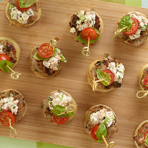 Tasty turkey sliders topped with savory turkey cream cheese and crumbled feta served on a wooden board, atop a colorful tablecloth.