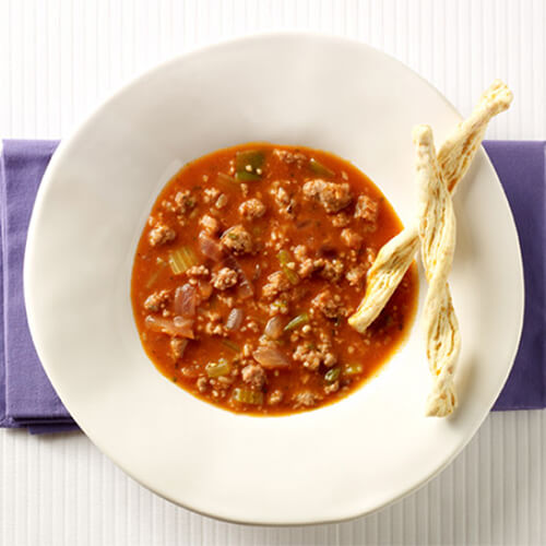 A savory tomato soup simmered with rice, bell peppers, celery, garlic and lean turkey breast, seasoned with spices. garnished with mini breadsticks in a white bowl.