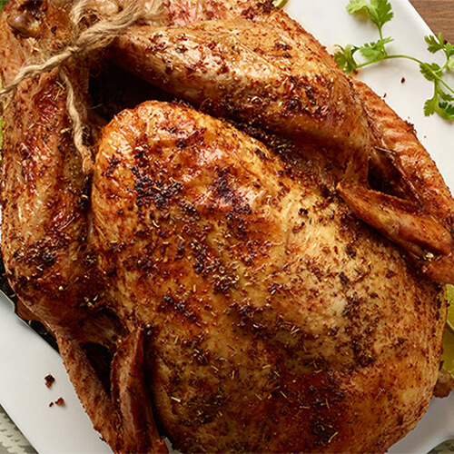 A whole turkey, made with a red pepper rub, served on a white plate garnished with sprigs of herbs and lime slices, atop a gray tablecloth.