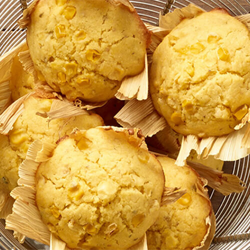 Cornmeal muffins featuring fresh whole kernel corn, cooked in cornhusks, served in a wired bowl atop a wooden table.