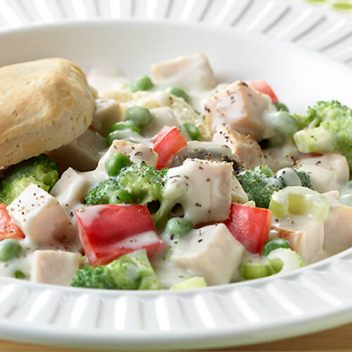 A dish made of cream, roasted turkey, and vegetables with a flaky biscuit all served in a white bowl on a wooden table.