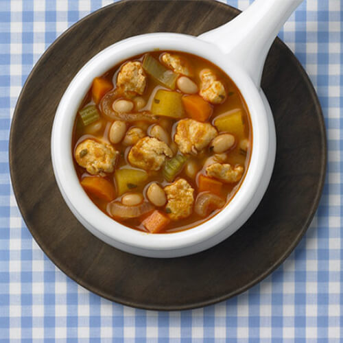 A filling amount of soup featuring a fair portion of turkey breast simmered with beans, rutabaga, carrots, and herbs, served in a white soup bowl atop a wooden plate, on a blue checkered tablecloth.