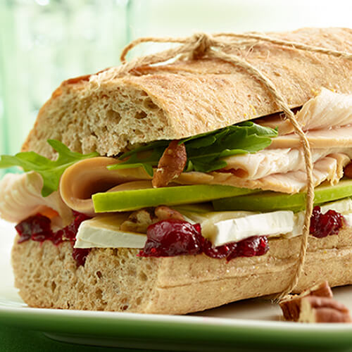 A sandwich filled with fresh arugula, thinly-sliced green apples, crunchy pecans, lean turkey breast, Brie cheese and swimming in a savory cranberry chutney, served on a white plate.