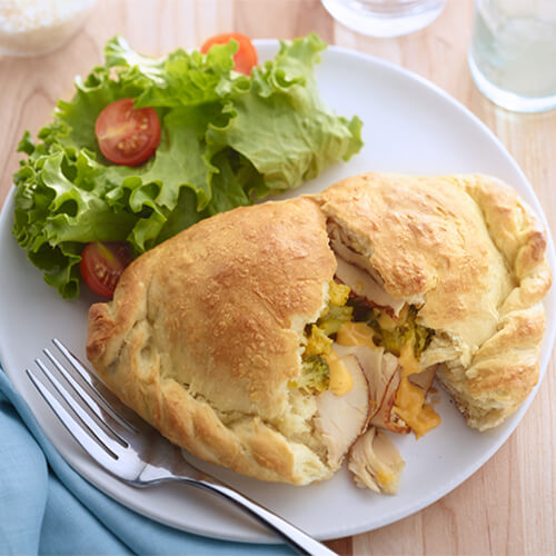A flaky calzone filled with a hearty portion of turkey, cheese, and broccoli florets, served with a side of a salad made from lettuce and tomato.