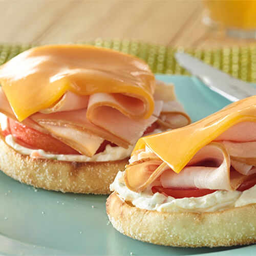 Toasted english muffins topped with cream cheese, tomatoes and turkey ham, on a blue plate atop a green table mat.