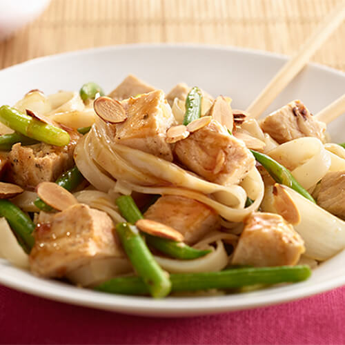 Stir fry made with a healthy portion of turkey, green beans, almonds and rice noodles served in a white bowl atop a ruby red cloth.