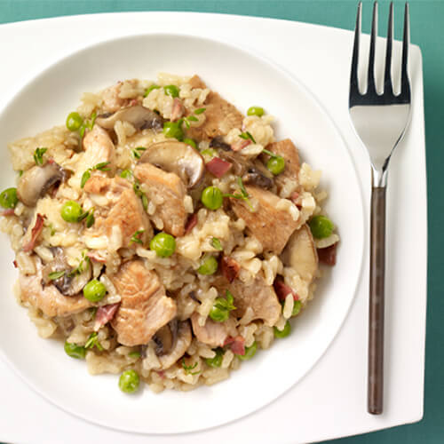 A generous portion of arborio rice served with pieces of turkey, bacon, mushrooms, and peas, made with a reduced white sauce, served in a white bowl, atop a cyan placemat.