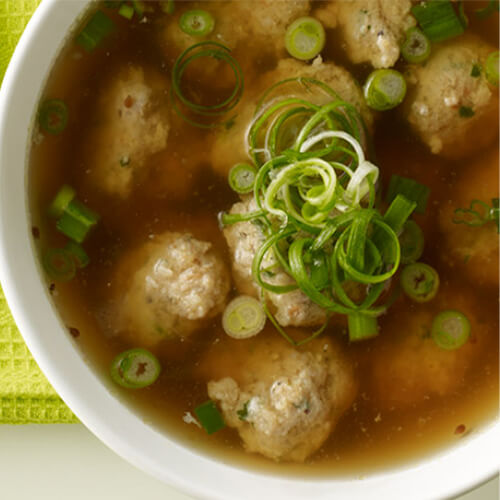 A savory soup with ground turkey, shrimp, cilantro and garlic dumplings simmered in a flavorful hoisin and lemongrass broth, served with a side of chopped green onions, on a white bowl, on a green cloth.