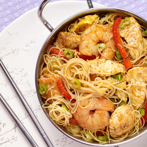 A hearty portion of rice stick noodles, ginger, curry, turkey, shrimp, and veggies, in a metal bowl, served on an asian-style plate, with a lavender woven tablecloth.