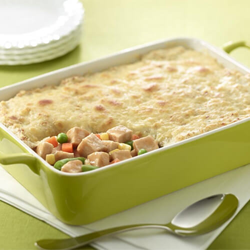 A hearty bake filled with loaded mashed potatoes, roasted turkey breast, and veggies served in a green casserole tray on a white cloth.