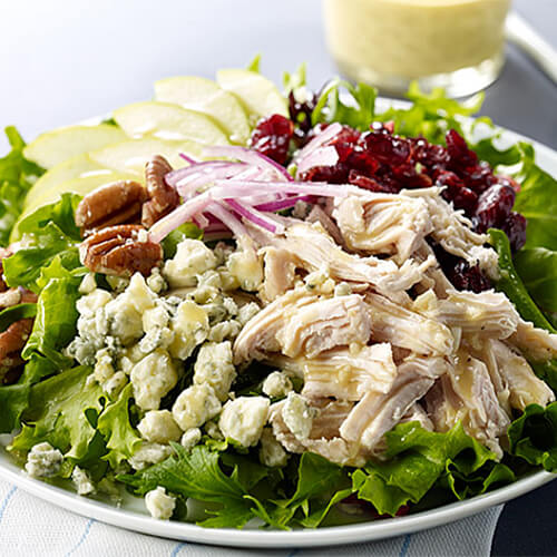 Roast turkey breast, cranberries, green apple, pecans, and blue cheese, in a bed of romaine, served with a mustard dressing on a white plate.