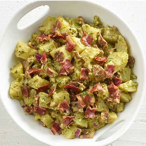 A hearty potato salad made with turkey bacon, guacamole, and jalpaneo served in a white bowl atop a wooden table.