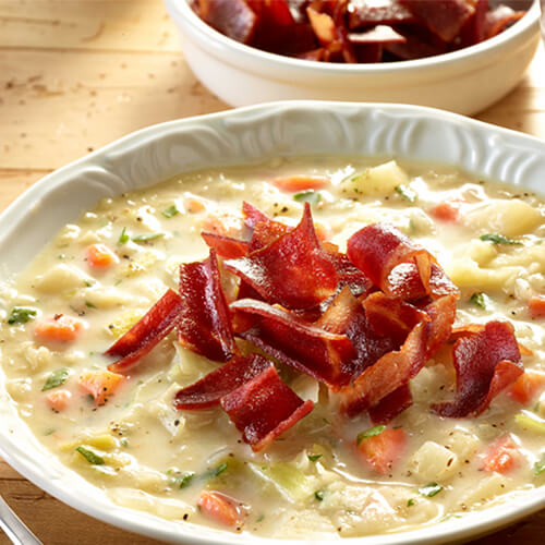 A soup made with potatoes, cabbage, parsley, carrots, and onion, topped with cut turkey bacon in a white bowl with a wooden background.
