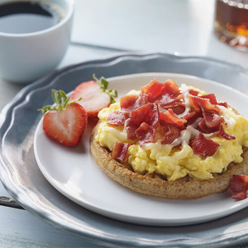 A whole grain waffle, topped with eggs, turkey bacon and melted cheese in a white plate, served with a cup of coffee and maple syrup.