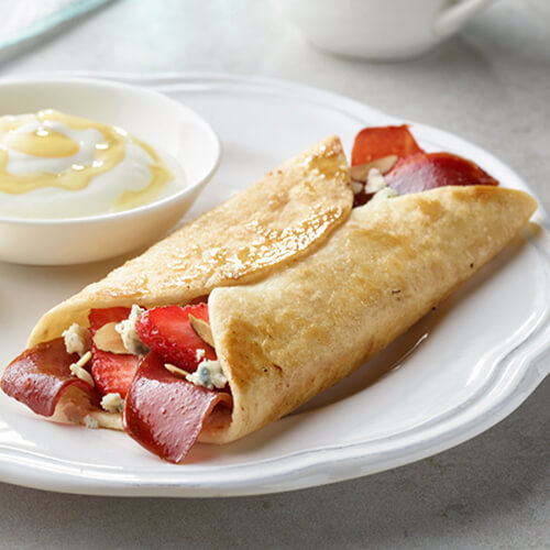 Fresh strawberries, crumbled blue cheese, and crunchy almonds and turkey bacon combined into a wrap, served with yogurt and honey on a white plate on a gray table.