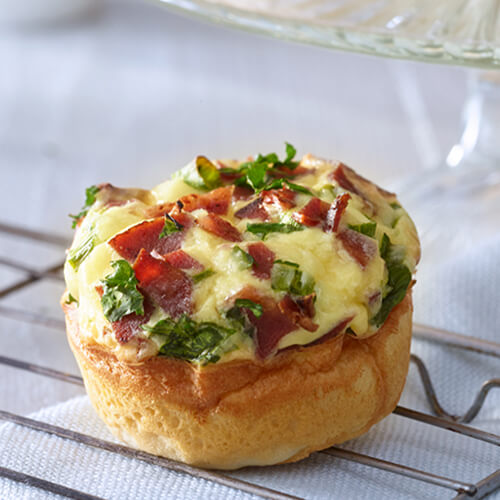 A flaky biscuit topped with a mixture of cream cheese and Swiss cheese, green onion, parsley, and JENNIE-O® Turkey Bacon on a baking rack with a white background.