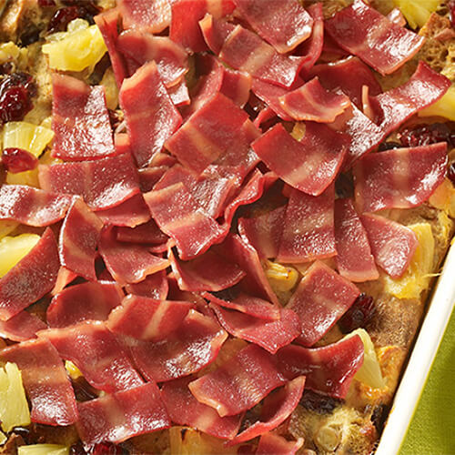 A hearty casserole made with cubed bread, cranberries, and pineapple tidbits, topped with a generous portion of turkey bacon and maple syrup, served in a green and white casserole tray.