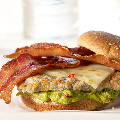 A delicious turkey burger topped with crisp turkey bacon, pepper Jack cheese and guacamole, served on white parchment paper.