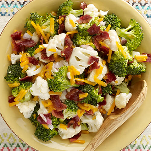 A summer salad filled with broccoli, cauliflower, cheese and onion, served on a large bowl atop a painted countertop.