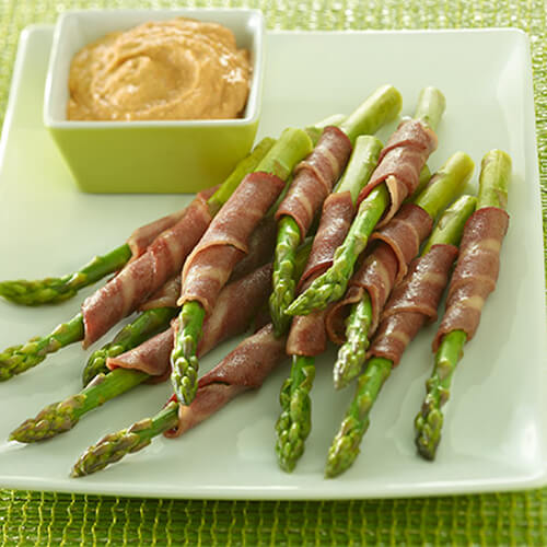 Grilled asparagus wrapped with crispy turkey bacon with a side of red pepper hummus, on a white plate atop a green woven tablecloth.