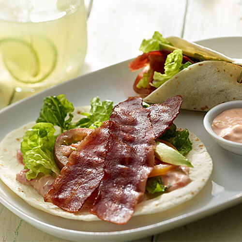 A tortilla filled with lettuce, tomato, avocado slices and turkey bacon, served with a side of salsa mayo on a white plate.