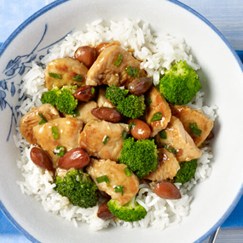 A hearty portion of white rice filled with broccoli and turkey tenderloin, and topped with a savory sauce, almonds, and chives, served in a porcelain bowl, atop a blue napkin.