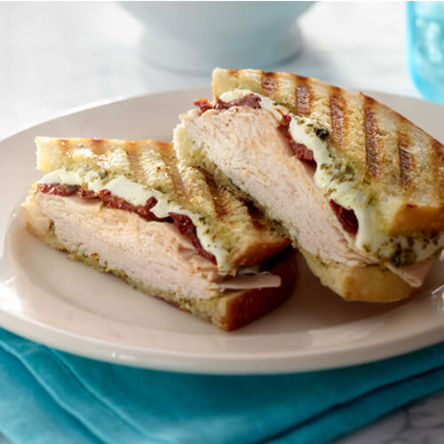 A halved grilled panini filled with browned turkey breast, sun-dried tomatoes and mozzarella cheese, served with pesto and mayo, on a white plate atop a sky blue cloth.