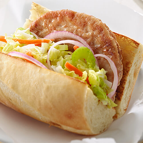 A turkey burger topped with cabbage, carrots, celery and onions, and a savory gravy, on a baguette roll, on parchment paper.