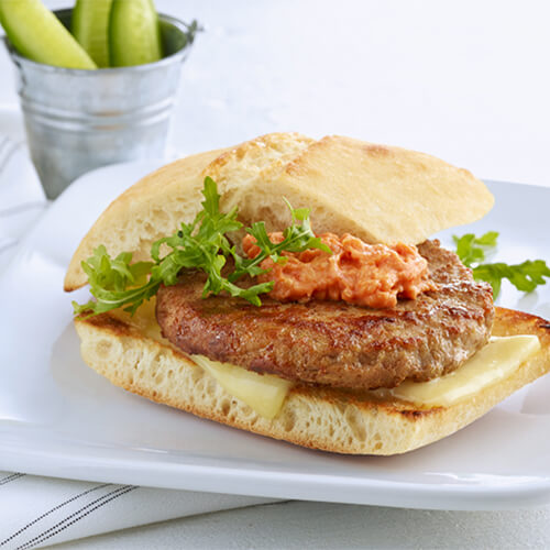 A juicy turkey burger topped with arugula, white cheddar cheese and a sundried tomato mayo, served on a white plate with a small serving bucket of pickles.