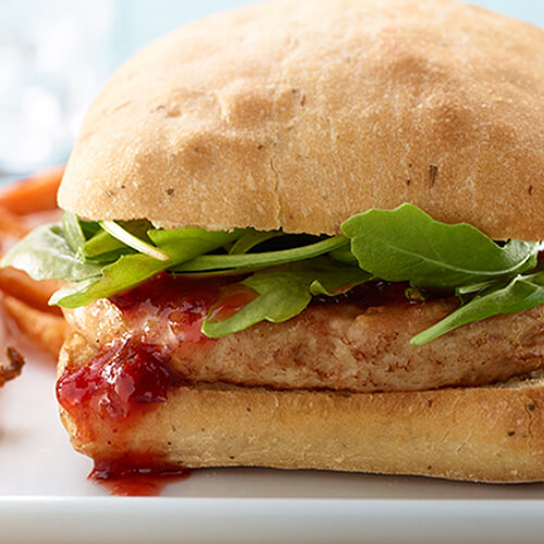 A perfect turkey burger topped with arugula and topped with a spicy citrus cranberry sauce, served on a white plate with a side of sweet potato fries.