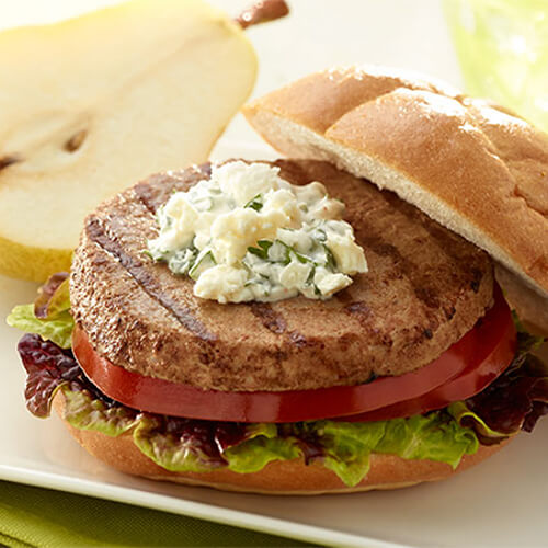 A turkey burger topped with Greek-style yogurt, lettuce, tomato, on a Kaiser roll, with a half pear, served on a white tray atop a green cloth.