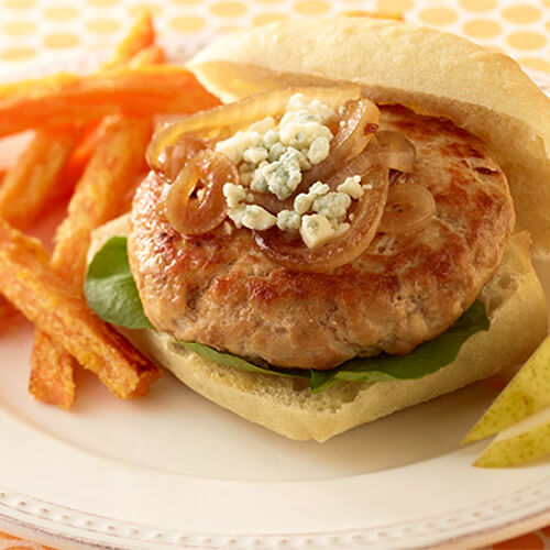 A delicious turkey burger, topped with Grilled onion, garlic, lettuce, and Worcestershire sauce, served with pear slices and sweet potato fries, on a white plate with a yellow patterned tablecloth.