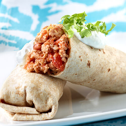 A burrito made with spicy salsa, beans, and lean ground turkey, topped with cilantro and sour cream, served with a side of tomatoes on a white plate, atop a blue floral tablecloth.