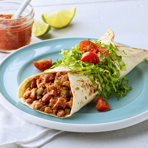 A hearty burrito filled with, JENNIE-O® ground turkey, HORMEL® turkey chili, topped with vegetables on a blue plate, with a side of salsa in a glass cup and lime wedges.