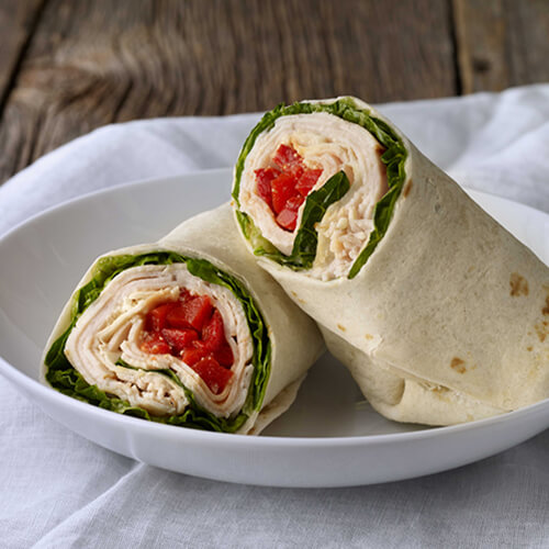 Turkey wraps filled with red peppers, lettuce, parmesan cheese and caesar dressing in a white bowl, under a white napkin and a wooden background.