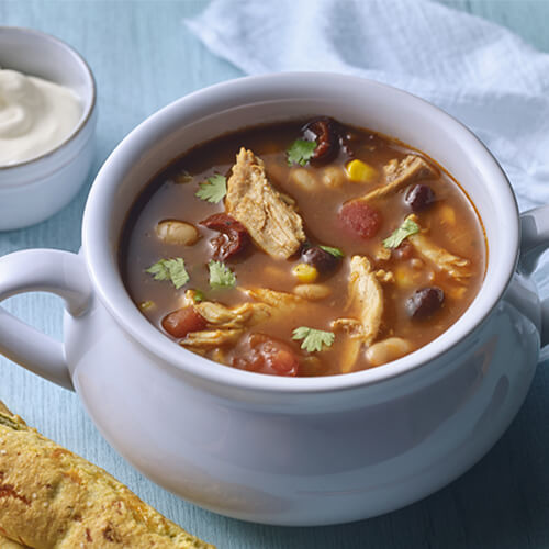 Turkey, cumin, salsa, corn, and two types of beans served in a lavender soup bowl, with a side of sour cream and breadsticks.