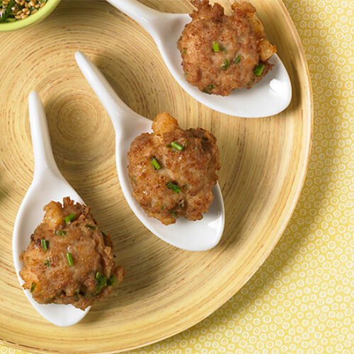 3 cakes made out of succulent turkey and shrimp, filled with chives, and jalapeno, on 3 spoons, with a side of a sesame dipping sauce, garnished with lime and cilantro, served on a wooden plate atop a yellow tablecloth.