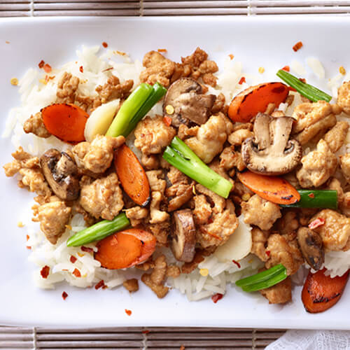 A classic chop suey made with extra lean ground turkey and filled with veggies, served on a bed of rice atop a white plate and a bamboo table mat, served with a side of sweet chili sauce.