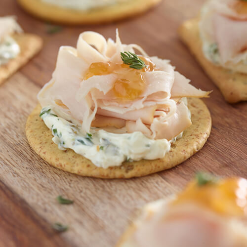 Crackers topped with soft herbed cream cheese, JENNIE-O® turkey, and a light amount of preserves, on a wooden cutting board.