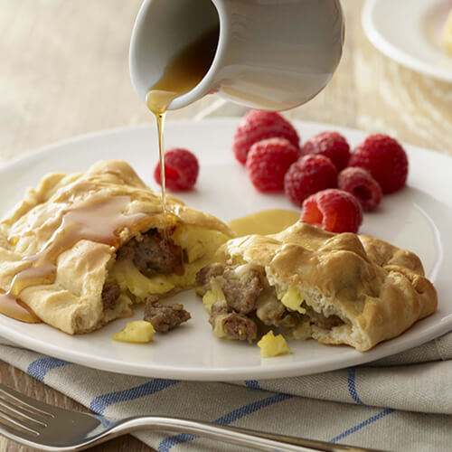 Flaky little pockets filled with eggs, cheese and turkey sausage, topped with maple apple syrup, served with a side of raspberries of on a white plate, atop a blue and white napkin on a wooden table.
