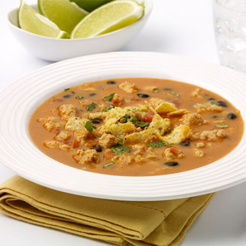 A hearty soup enchilada soup served with crushed tortilla chips, beans, ground turkey, and vegetables, served in a white bowl, on a yellow napkin.