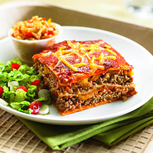 A hearty portion of ground turkey, sautéed onions, shredded cheese and spicy enchilada sauce served in a casserole-style, served with a side of salad on a white plate atop a green cloth.