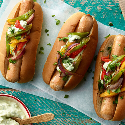 Three fajita turkey dogs topped with bell peppers and onions on a white background.