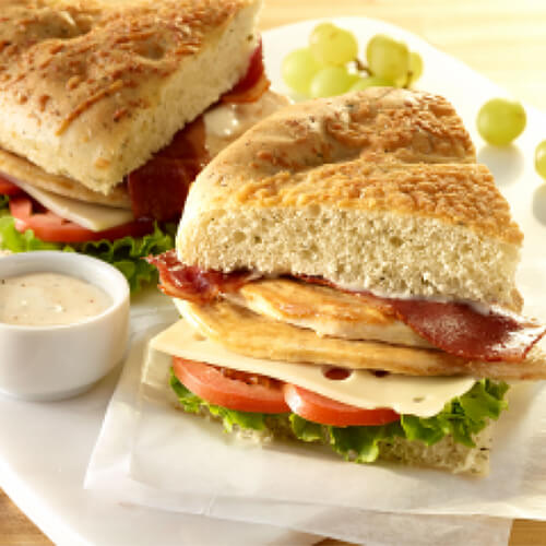 A flaky focaccia sandwich filled with layers of fresh veggies, turkey breasts, and Swiss cheese served with a bold parmesan dressing on a white cutting board on a wooden table.