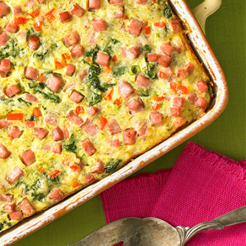 An egg casserole filled with spinach, Swiss cheese, green onions, red bell pepper, and JENNIE-O® turkey ham, topped with lemon slices and dill weed, in an orange casserole tray.