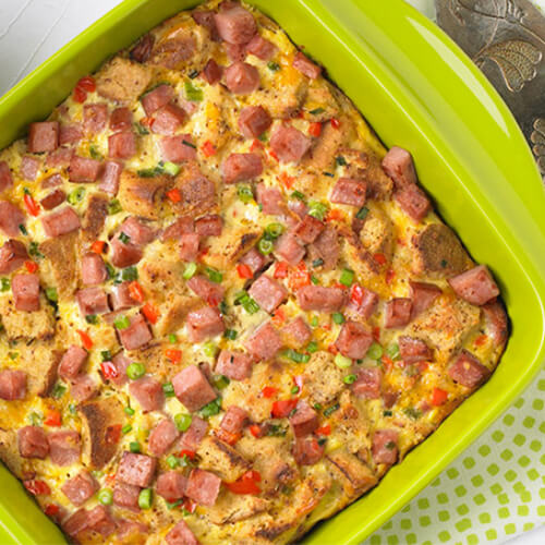 A hearty portion of lean turkey ham, eggs, bell peppers, onions, and sharp cheddar, served in a lime green baking dish, served on a patterned cloth.