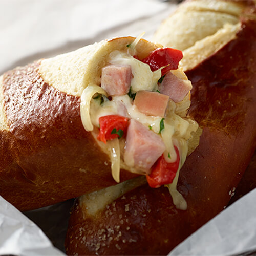Turkey ham, onion, peppers, parsley, thyme, and cheese, served in a pretzel roll in a wired bowl lined in parchment paper.